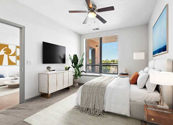 Bedroom at 7001 Arlington at Bethesda in Bethesda, MD, featuring a bed, ceiling fan, wall-mounted TV, large window, and view into the living area.