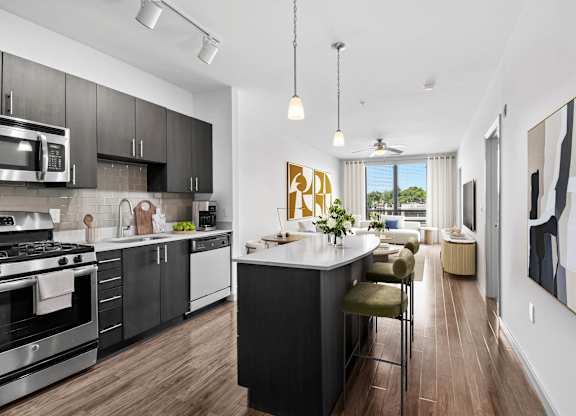 Open-concept kitchen and living area at 7001 Arlington at Bethesda in Bethesda, MD, with an island, espresso cabinetry, stainless-steel appliances, wood-style flooring, and large windows.