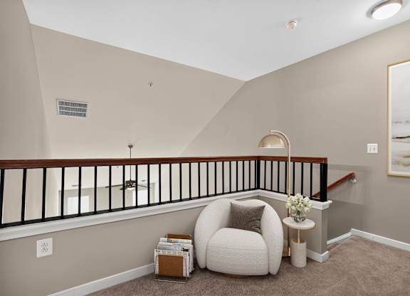 Virtually staged open loft at Enclave at Emerson overlooking the main living area, with neutral carpeting, a rounded lounge chair, side table and floor lamp.