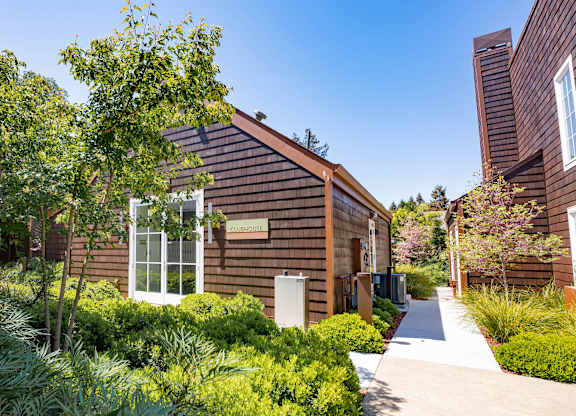 A small building with a brown roof and a white door is surrounded by green plants.