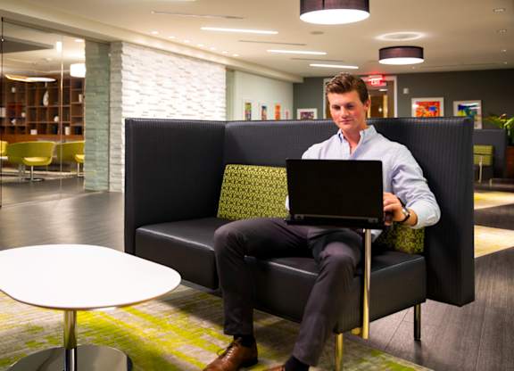 A man in a white shirt and black pants is sitting on a black couch with a laptop on his lap.