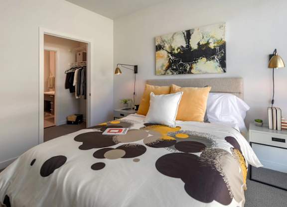 A bed with a white comforter and a brown and black pattern is in the foreground of a room with a painting on the wall and a clothing rack in the background.