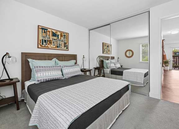 Bedroom with wood bed frame, nightstand, lamp, and window with blinds at Old Orchard Apartments
