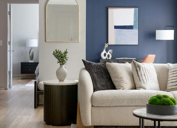 A living room with a white couch, a round table with a plant on it, and a mirror on the wall.