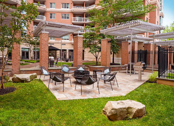 A patio with chairs and a table is surrounded by a grassy area and a building in the background.