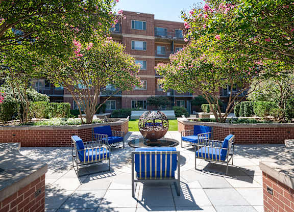 Courtyard seating area with four blue striped chairs around a round metal fire feature, framed by trees and brick buildings.