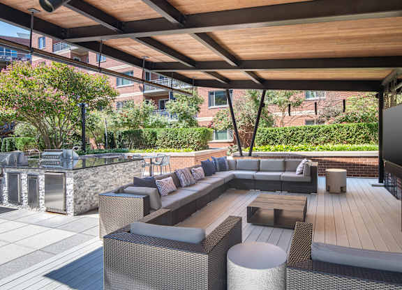 Covered outdoor lounge with gray sectional seating, white side tables, a television and nearby grilling stations.
