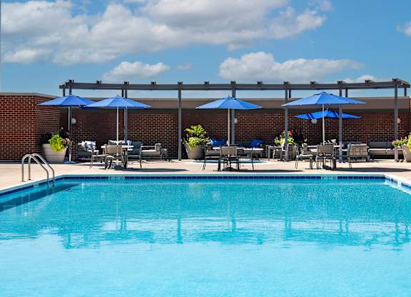 Low-angle view across the outdoor swimming pool toward blue umbrellas, tables, lounge chairs and the sundeck.