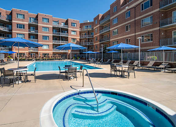 Round outdoor spa beside the Vantage Mosaic swimming pool, with tables, blue umbrellas and brick apartment buildings.