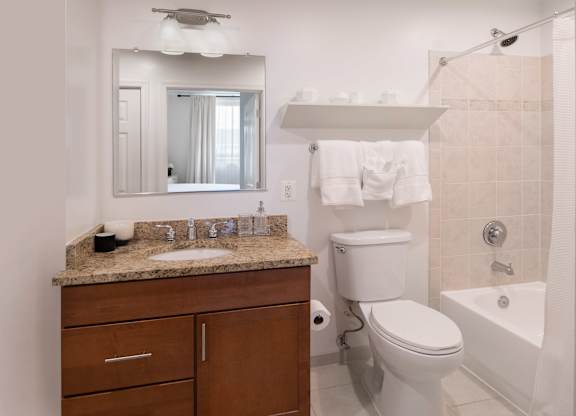 Model apartment bathroom with a granite vanity, wood cabinet, mirror, toilet and white tub with tile surround.