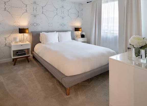 Model bedroom with a white bed, gray upholstered frame, matching nightstands, black lamps and a light patterned accent wall.