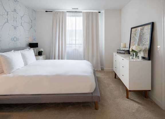 Bright model bedroom with a white bed, gray upholstered frame, white dresser, floor-length curtains and patterned accent wall.