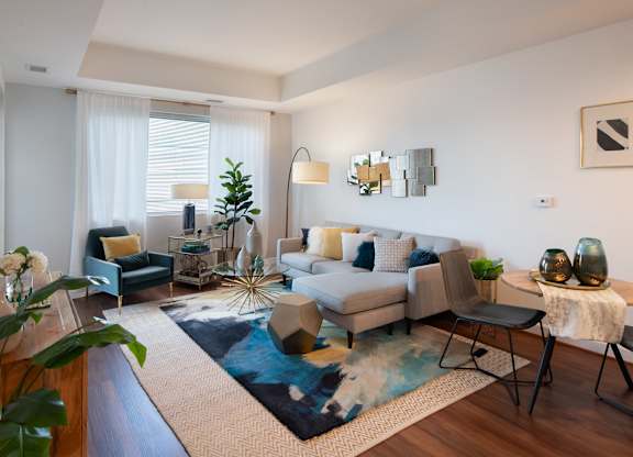 Sunlit model living room with a gray sectional, blue area rug, glass coffee table, accent chair and large windows.