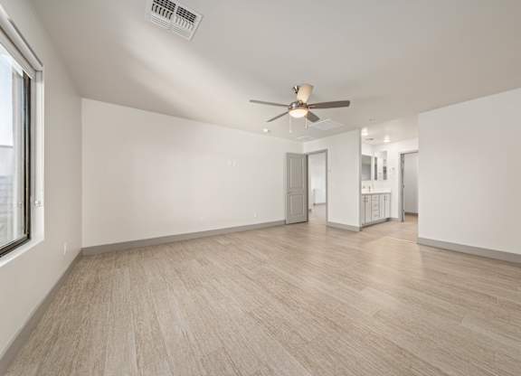 A spacious room with a ceiling fan and light, hardwood flooring, and a window on the left side.
