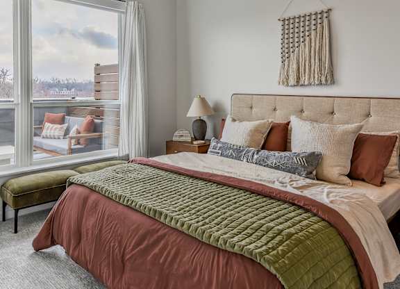 A bed with a green comforter and a red blanket is in the foreground of a room with a window showing a view of a snowy landscape.