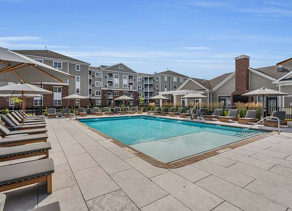 A large swimming pool surrounded by lounge chairs and umbrellas in front of apartment buildings.