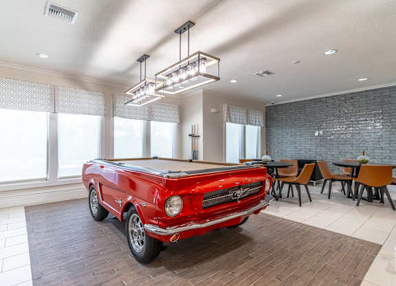 A red vintage car is parked in a spacious room with a dining table and chairs.