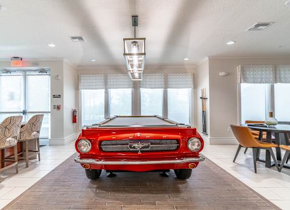 A red vintage car is parked in a spacious room with a dining table and chairs.
