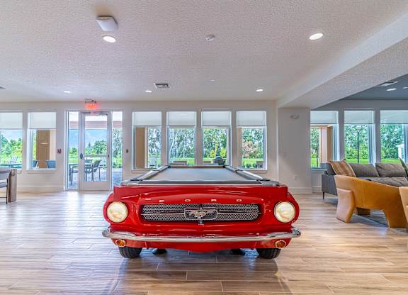 A red vintage car is parked in a spacious room with wooden floors and large windows.