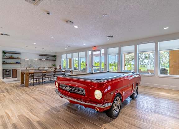 A red vintage car is parked in a spacious room with wood flooring and large windows.