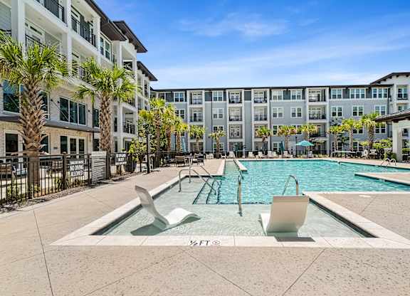 A swimming pool with two loungers in front of a building with palm trees.