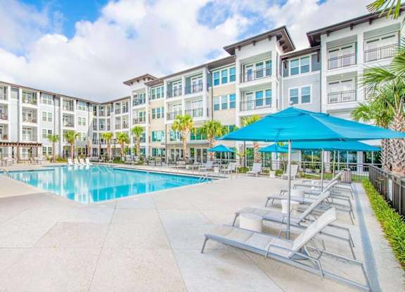 A large swimming pool surrounded by sun loungers and umbrellas in front of apartment buildings.