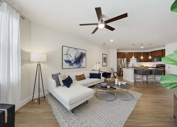 Open living area with a sectional, ceiling fan, and wood-tone flooring extending to the kitchen island and bar stools.