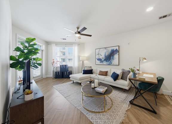 Living room with a sectional, area rug, corner windows with blinds, and a writing desk set against the far wall.