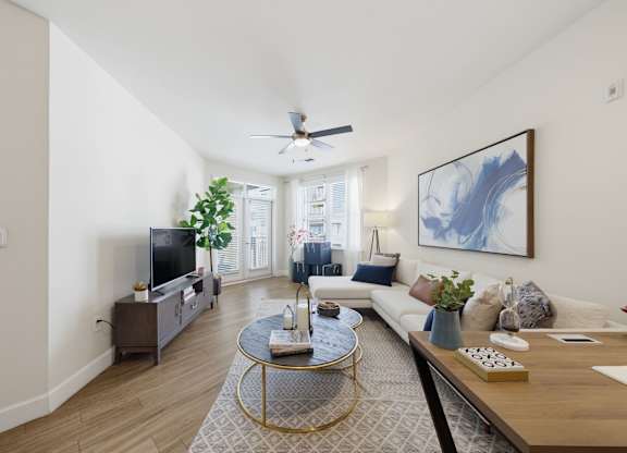 Living area with wood-tone flooring, a ceiling fan, media console, and a shuttered balcony door letting in daylight.