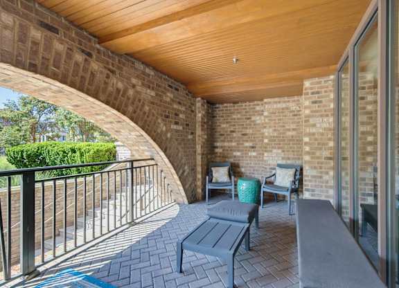 Covered terrace under a brick archway, with a wood-slat ceiling, two chairs, a chaise, and a railing over the garden.
