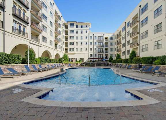 Pool with a bubbling shallow entry, handrails, and loungers on both sides, enclosed by the apartment building.