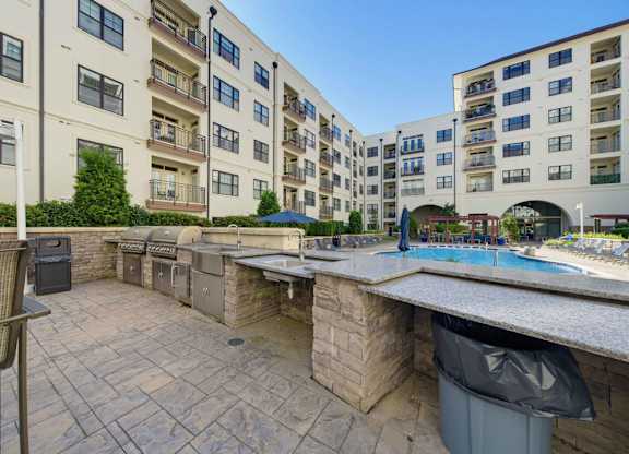 Outdoor grilling station with two gas grills, a prep sink, a stone bar counter, and umbrella tables beside the pool.