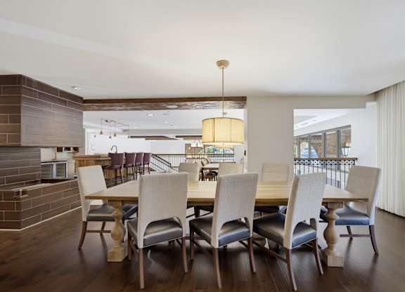 Long clubroom dining table seating ten under a drum pendant, with a tile fireplace on one side and a bar beyond.