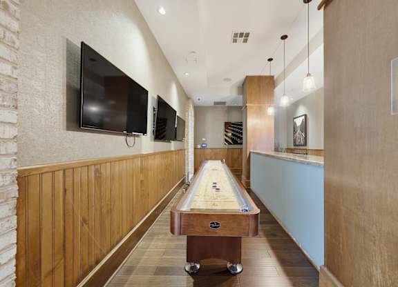 Game area with a shuffleboard table, three mounted screens, wine racks, and a bar counter along the panelled far wall.