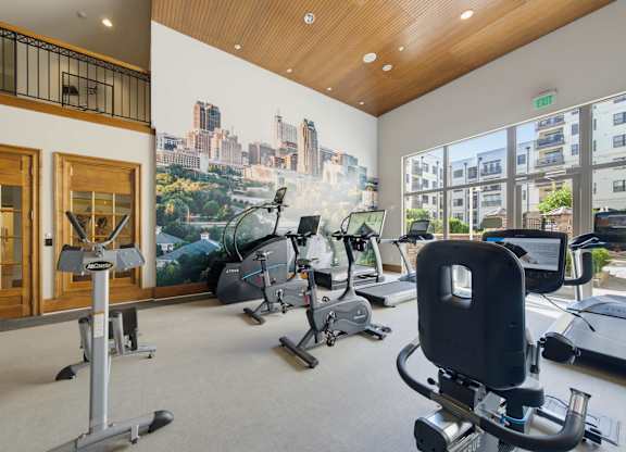Fitness room with studio bikes, a stair climber, and an abdominal machine in front of a Raleigh skyline mural.