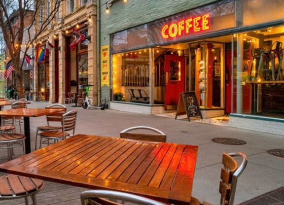 Sidewalk cafe tables and metal chairs outside a lit coffee shop, with string lights strung above the storefronts.