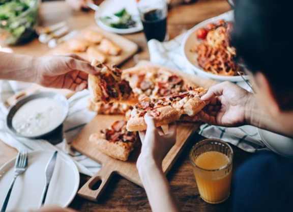 Overhead view of a shared meal on a wood table, with pizza on a wooden board, a salad bowl, and drinks alongside.