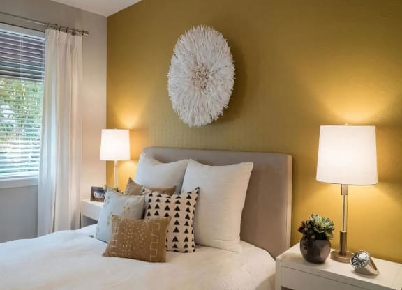 Bedroom with an upholstered headboard against a gold accent wall, matching nightstand lamps, and a curtained window.