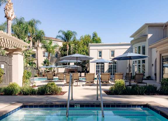 Pool at Amerige Pointe with entry steps and handrails, ringed by lounge chairs, umbrellas, and mature palm trees.