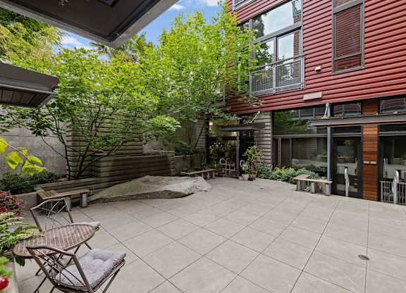 A patio with a table and chairs is surrounded by a red building and trees.