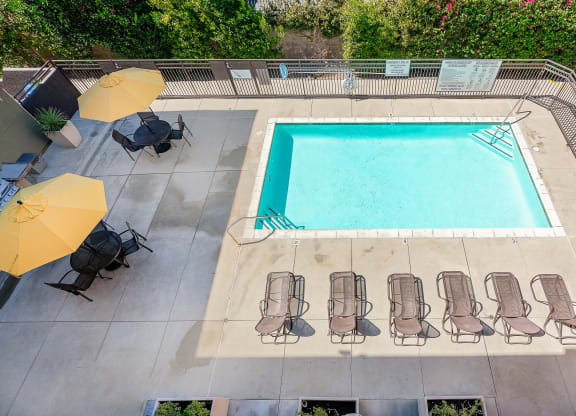 Apartment Rancho Cucamonga, CA - Villas at the Gardens - Elevated View Of Resort Style Pool With Sundeck Lounge Chairs, Yellow Umbrellas, And Patio Seating.
