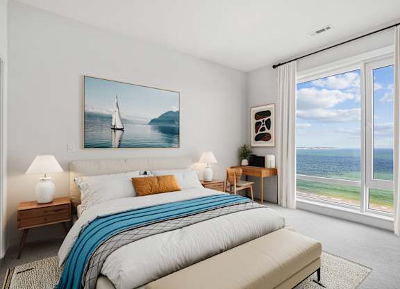 Carpeted penthouse bedroom with nightstands and a desk set beside a tall window over the sand and the open water.