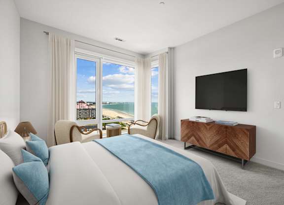 Carpeted penthouse primary bedroom with two chairs set at corner windows that look over the beach and the rooftops.