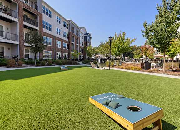 A cornhole board sits on a green lawn in front of apartment buildings.