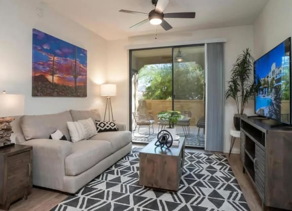 North Scottsdale AZ Apartments for Rent - Desert Parks Vista - Living Room with Wood Flooring, Black and White Rug, Beige/Grey Sofa, Wooden Coffee Table, Side Table with Lamp, Standing Lamp, Wooden TV Stand with Drawers, TV, House Plants, Sliding Glass Door to Balcony, and Ceiling Fan