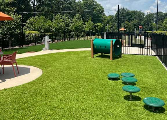 A green cylindrical object is in the middle of a grassy area with a red bench to the left.