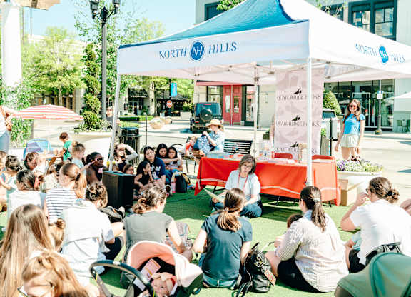 A group of people are sitting on the grass in front of a tent with the sign