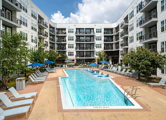 A large swimming pool surrounded by lounge chairs and umbrellas in front of apartment buildings.