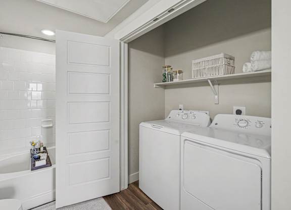 Laundry closet with a white top-load washer and dryer under a wall shelf, next to a bathroom with a tiled tub and shower.