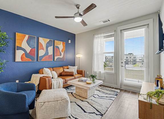 Living room with a blue accent wall, ceiling fan, wood-look floors, and a window beside a glass balcony door with blinds.